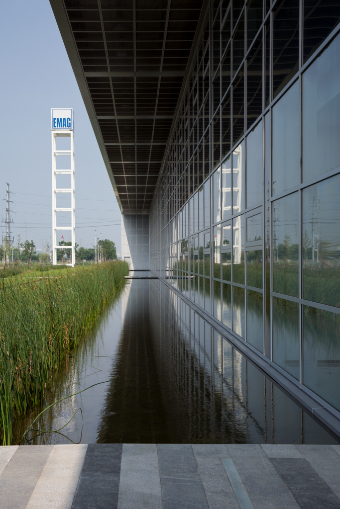 Eingang des Industriegebäudes mit strukturierter Fassade, auskragendem Dach und vorgelagerter Wasserfläche